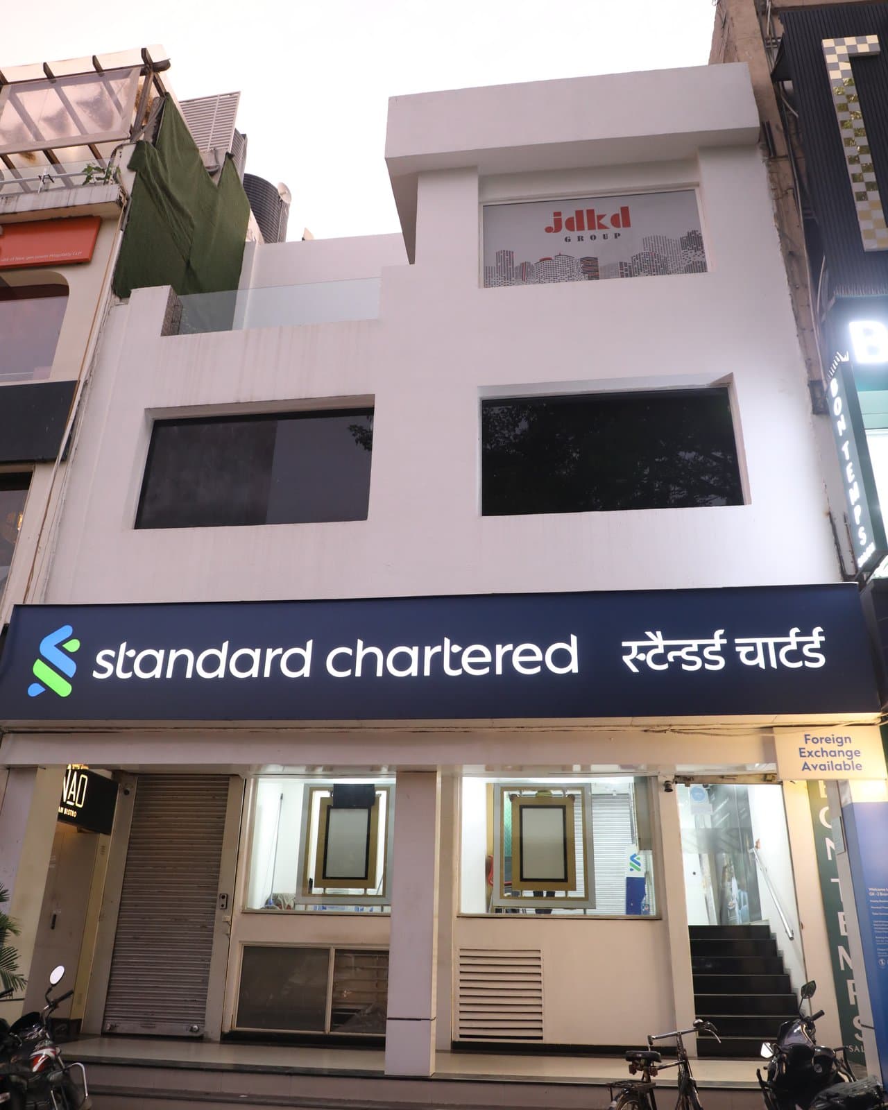 The full M-39 frontage at dusk: the JDKD mark above, the Standard Chartered branch below.